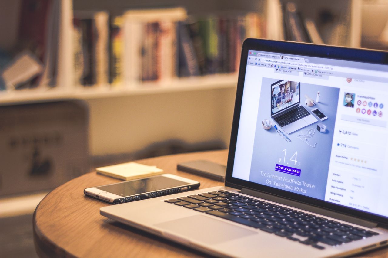 our-story A cozy modern workspace featuring a laptop displaying a website, a smartphone, and books on shelves.