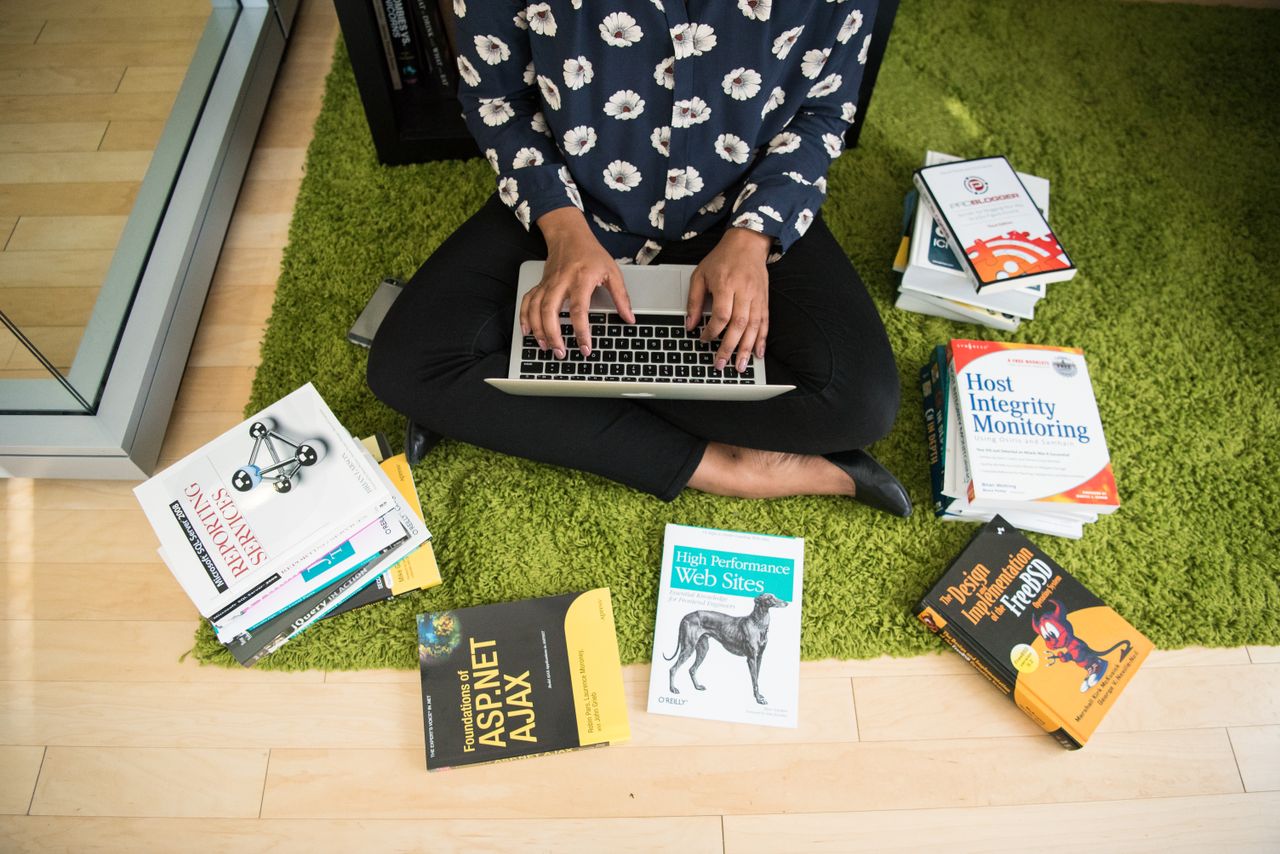 creative Woman sitting on green rug working on laptop, surrounded by technology books in a modern room.